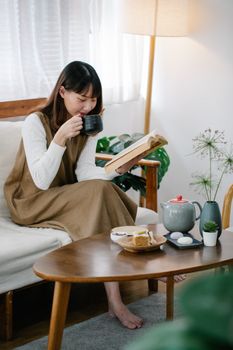 Adult woman reading a book while sipping tea in a cozy indoor setting.