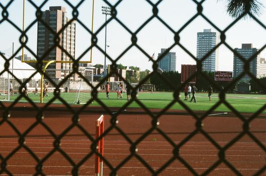 Soccer players on an urban field seen through a metal fence, with cityscape in the background.