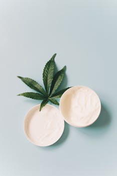 Top view of cannabis-infused skincare cream with hemp leaf on light blue background.