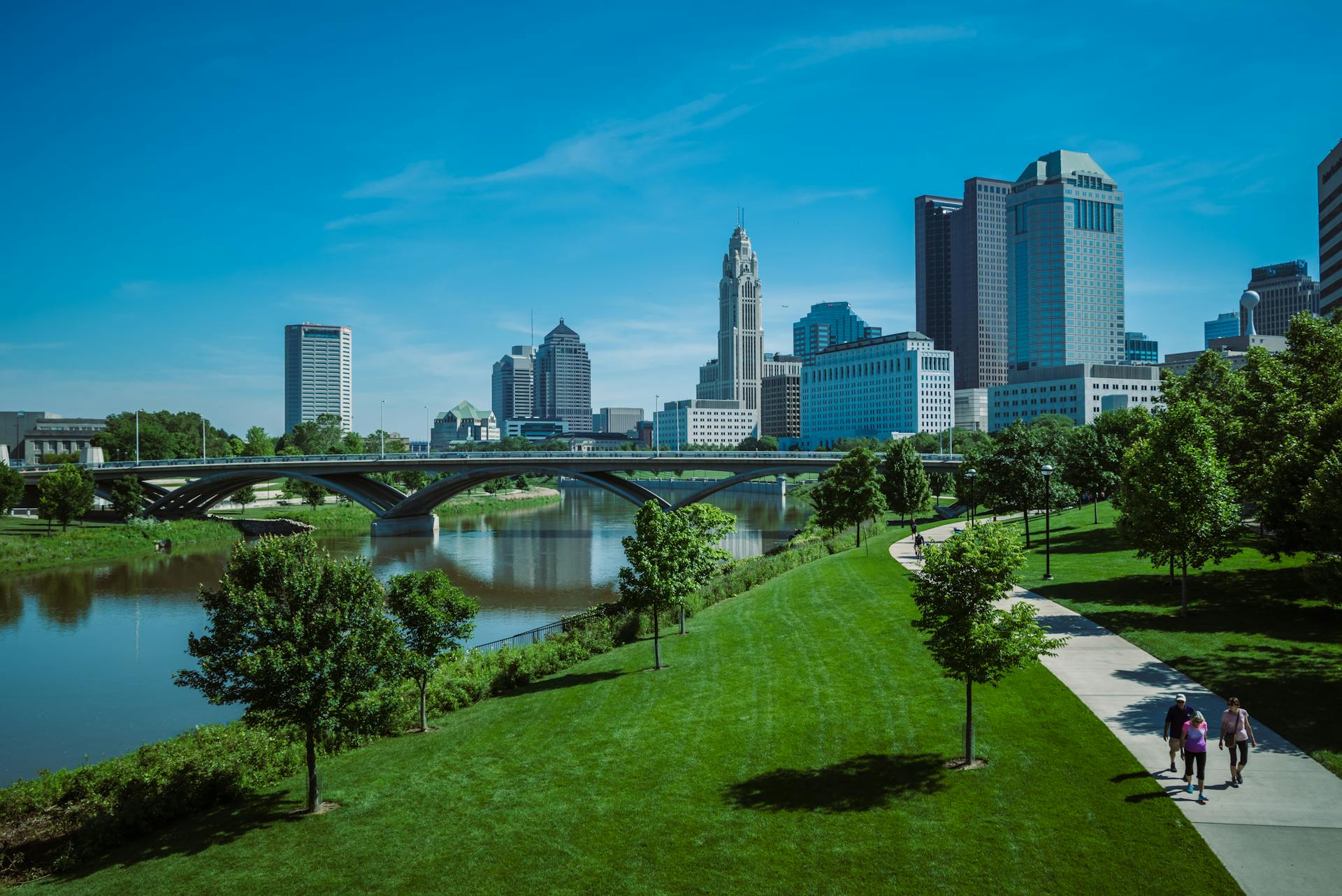 Sunny view of Columbus skyline with a park, bridge, and river in the foreground.