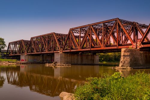 Free A rusted railway bridge with intricate design reflected in a calm river under clear skies. Stock Photo