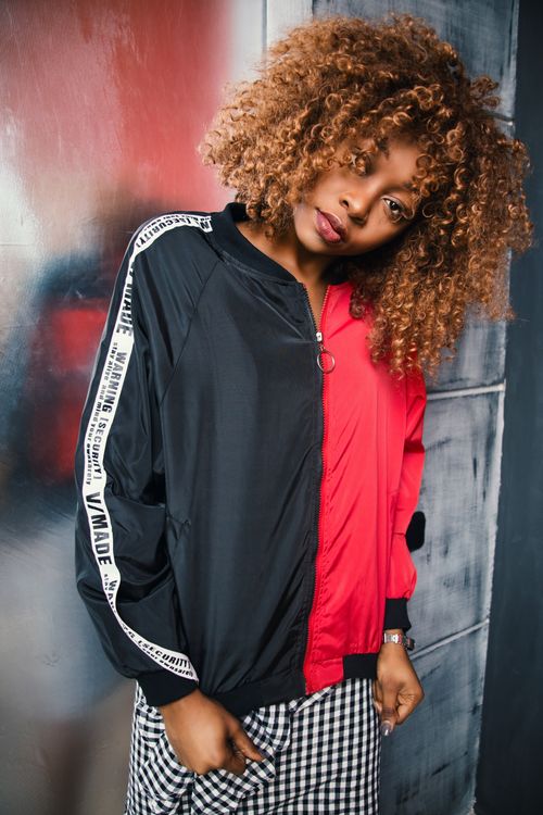 Free Black woman with curly hair wearing a fashionable red and black jacket posing indoors with a modern vibe. Stock Photo