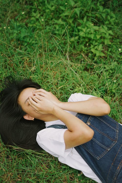 A Woman in White Shirt and Denim Jumper Lying on Green Grass while Covering Her Face