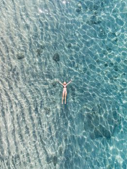 Aerial drone shot of a woman leisurely floating in a crystal clear blue sea, showcasing serenity and relaxation.