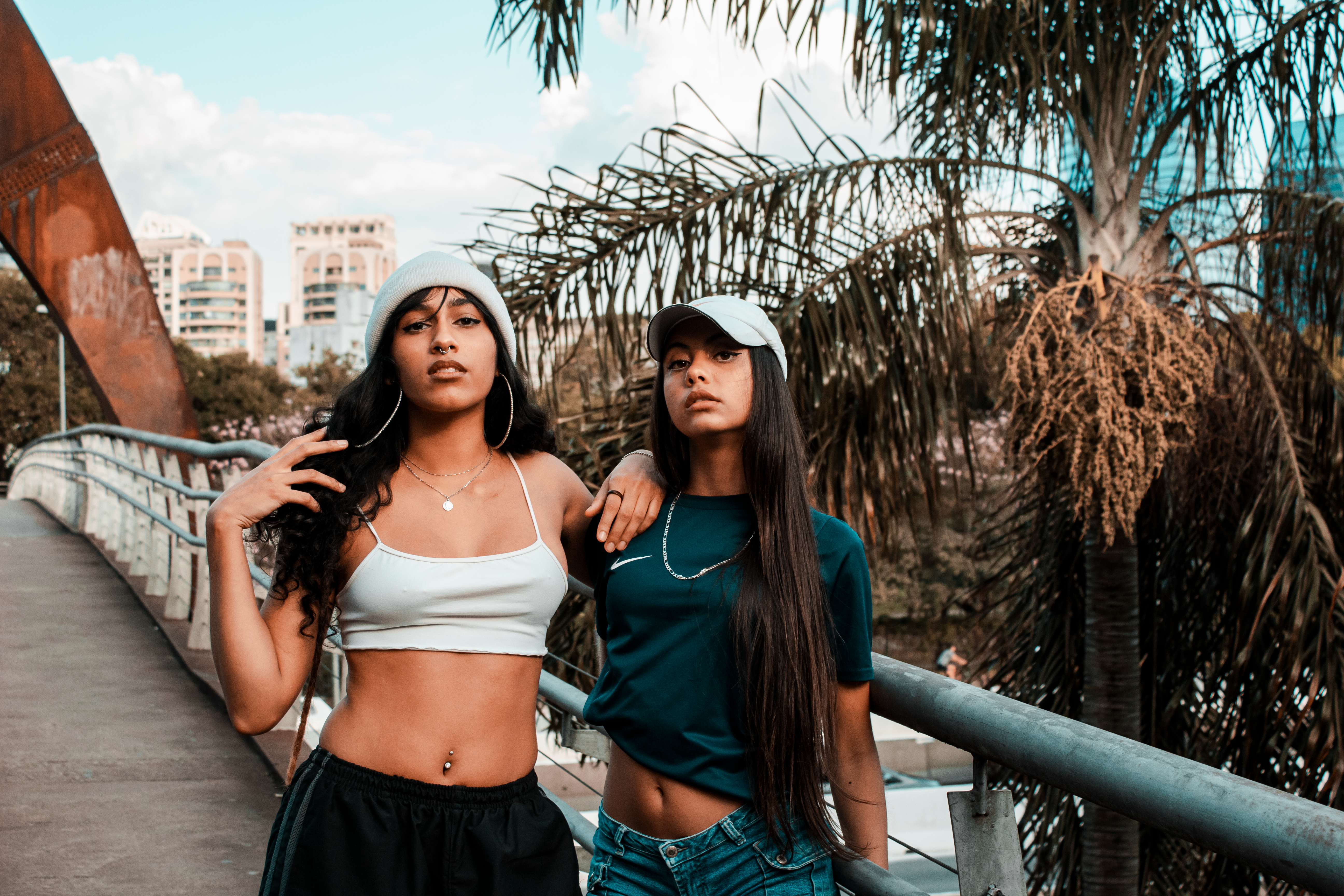 Two stylish women pose confidently on a bridge in an urban setting with palm trees.