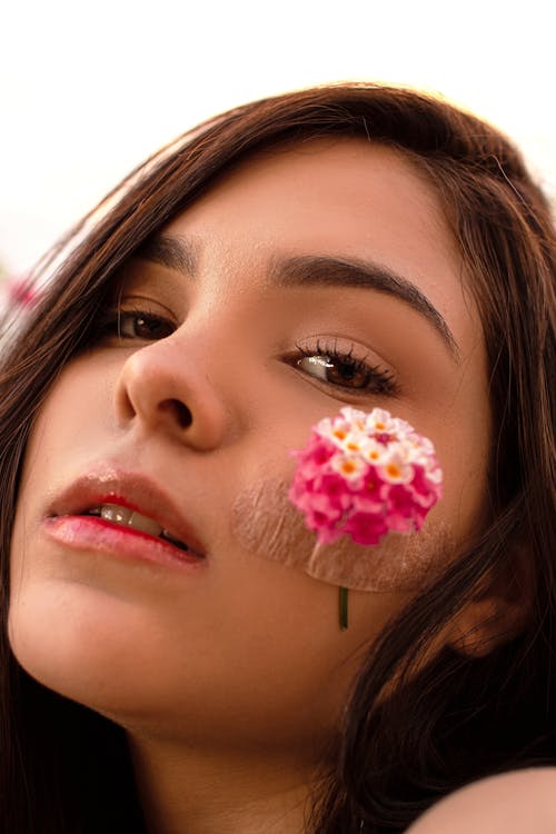 Headshot of charming young female with dark hair and colorful pink flower on cheek with band aid looking at camera