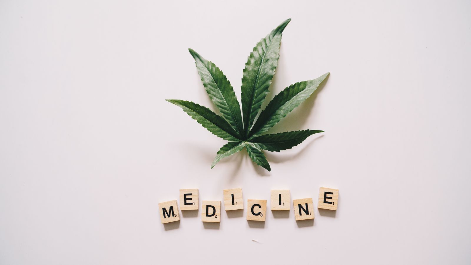 Flat lay of cannabis leaf and 'MEDICINE' spelled with Scrabble tiles on white background.