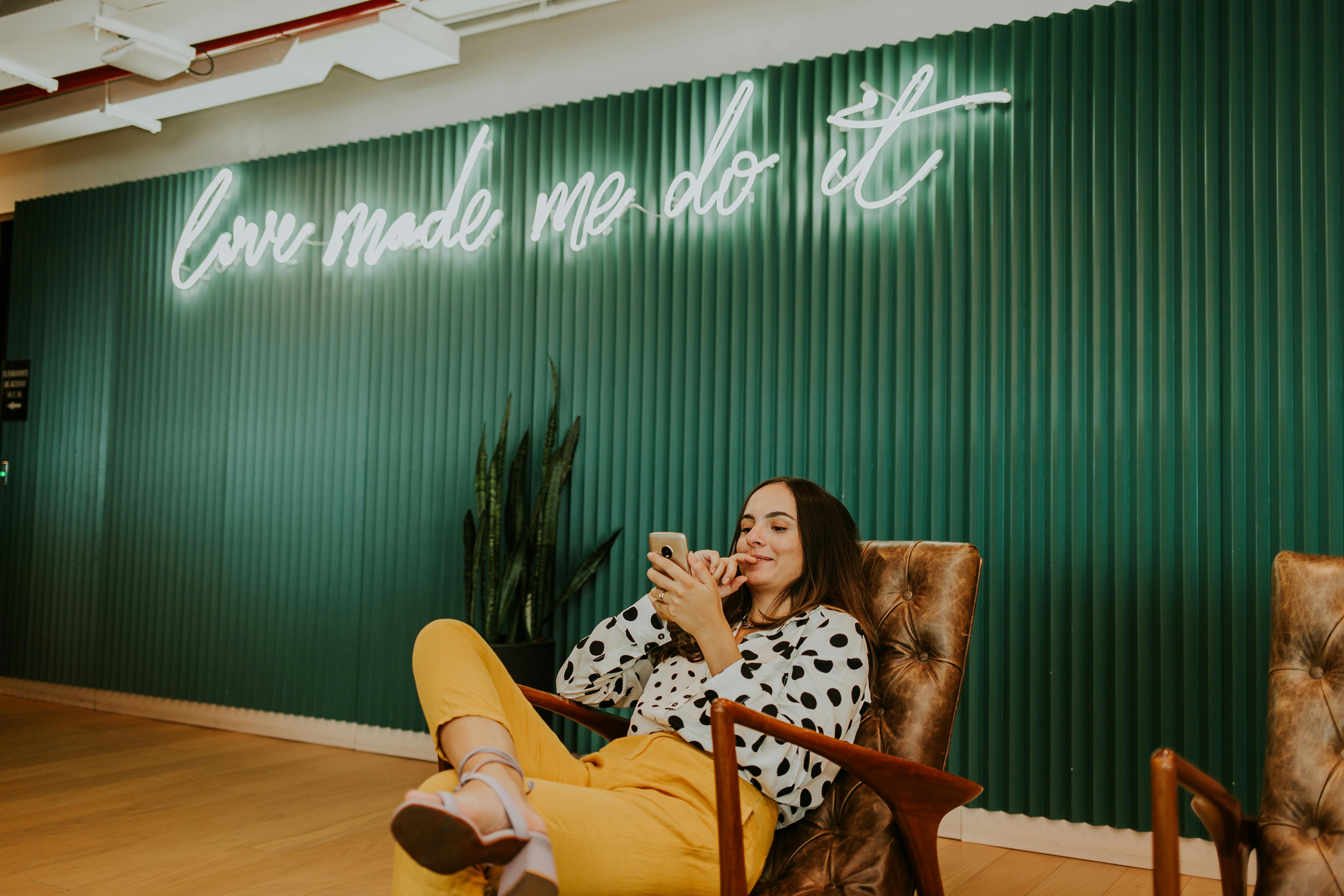 a woman in white and black polka dot shirt sitting on brown chair holding a cellphone