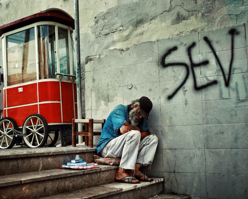 Un hombre mayor con una larga barba descansa junto a un carrito en una calle urbana, al lado de graffitis.