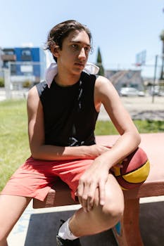 Teenage boy in sports attire resting with a basketball on an outdoor court bench.