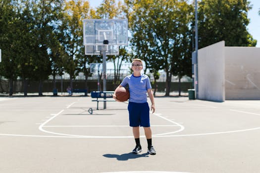 A cheerful boy in glasses holding a basketball on a sunny outdoor court.