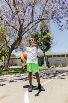 A young boy in bright shorts stands confidently with a basketball outdoors on a sunny day.