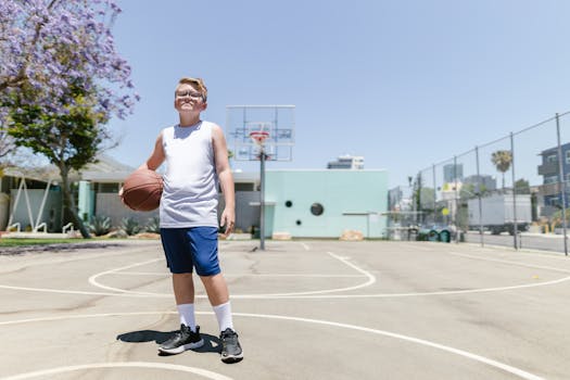 A young boy in blue shorts and a white tank top holding a basketball on an outdoor court under clear skies.