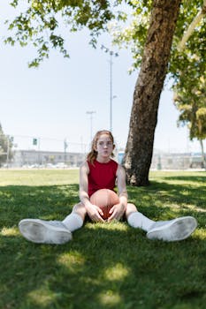 A young girl sitting under a tree in a park holding a basketball, enjoying a sunny day outdoors.