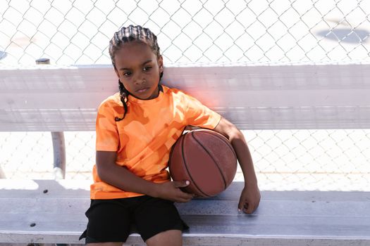 A young boy sitting with a basketball on an outdoor bench in casual attire.