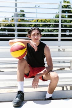 Teen sitting on outdoor bleachers holding a basketball, showcasing casual athletic wear.
