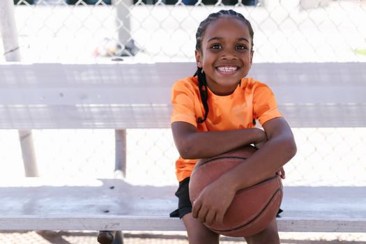 A young girl in an orange shirt, smiling, holding a basketball while sitting on a bench outdoors.