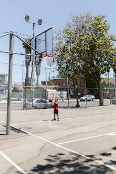 A young boy in a red shirt practices basketball shooting on an outdoor court under a clear sky.
