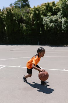 A young boy dribbling a basketball on an outdoor court, wearing an orange shirt.