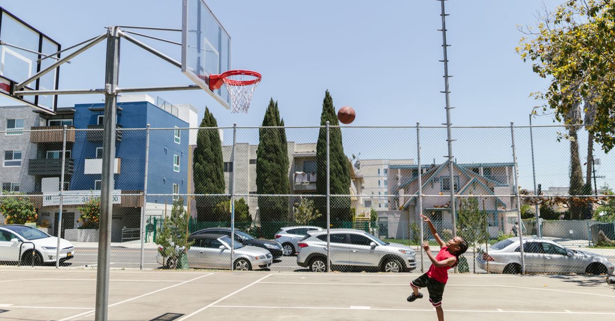 Kids from diverse backgrounds playing a pickup basketball game on a well-lit outdoor court in an urban neighborhood at golden hour