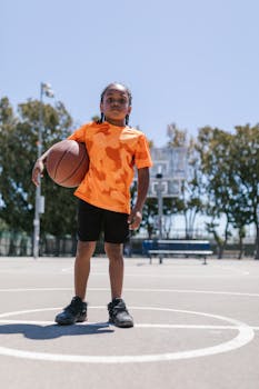 An African American boy with braided hair holding a basketball on an outdoor court.