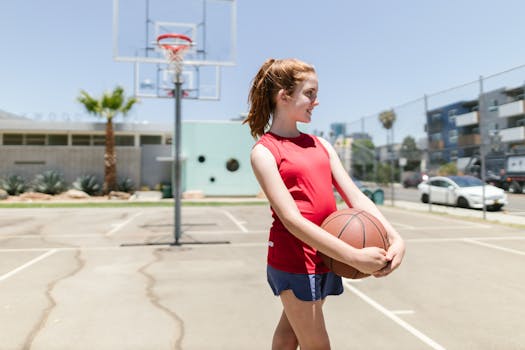A teenage girl in sportswear holding a basketball on an outdoor court on a sunny day.