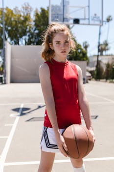 A young girl in a red jersey holding a basketball on an outdoor court during the day.