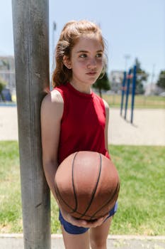 Redheaded girl in red tank top holds basketball outdoors on sunny day.