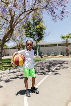 African American boy with braided hair holding basketball on court at park, outdoors, during daytime.
