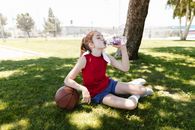Woman in Red Tank Top and Black Shorts Sitting on Green Grass Field Drinking from Clear