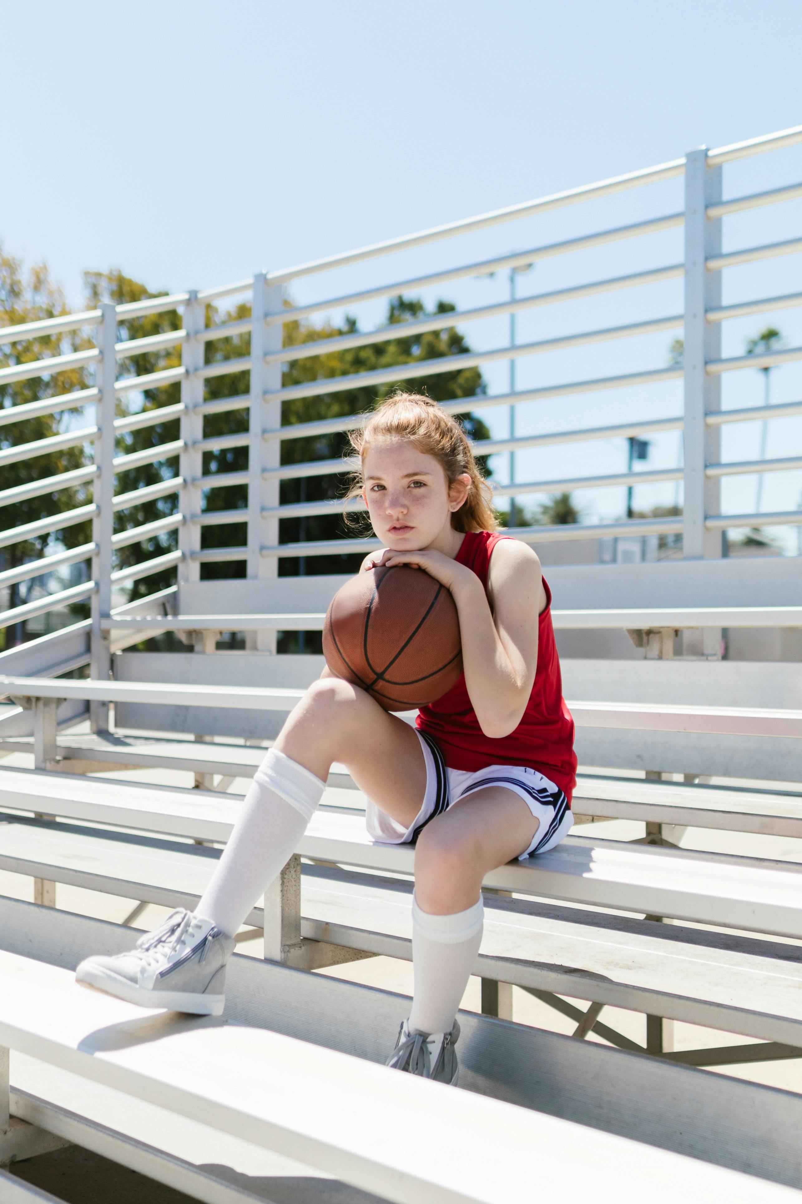 a young girl in red tank top holding a basketball while sitting on the bleachers