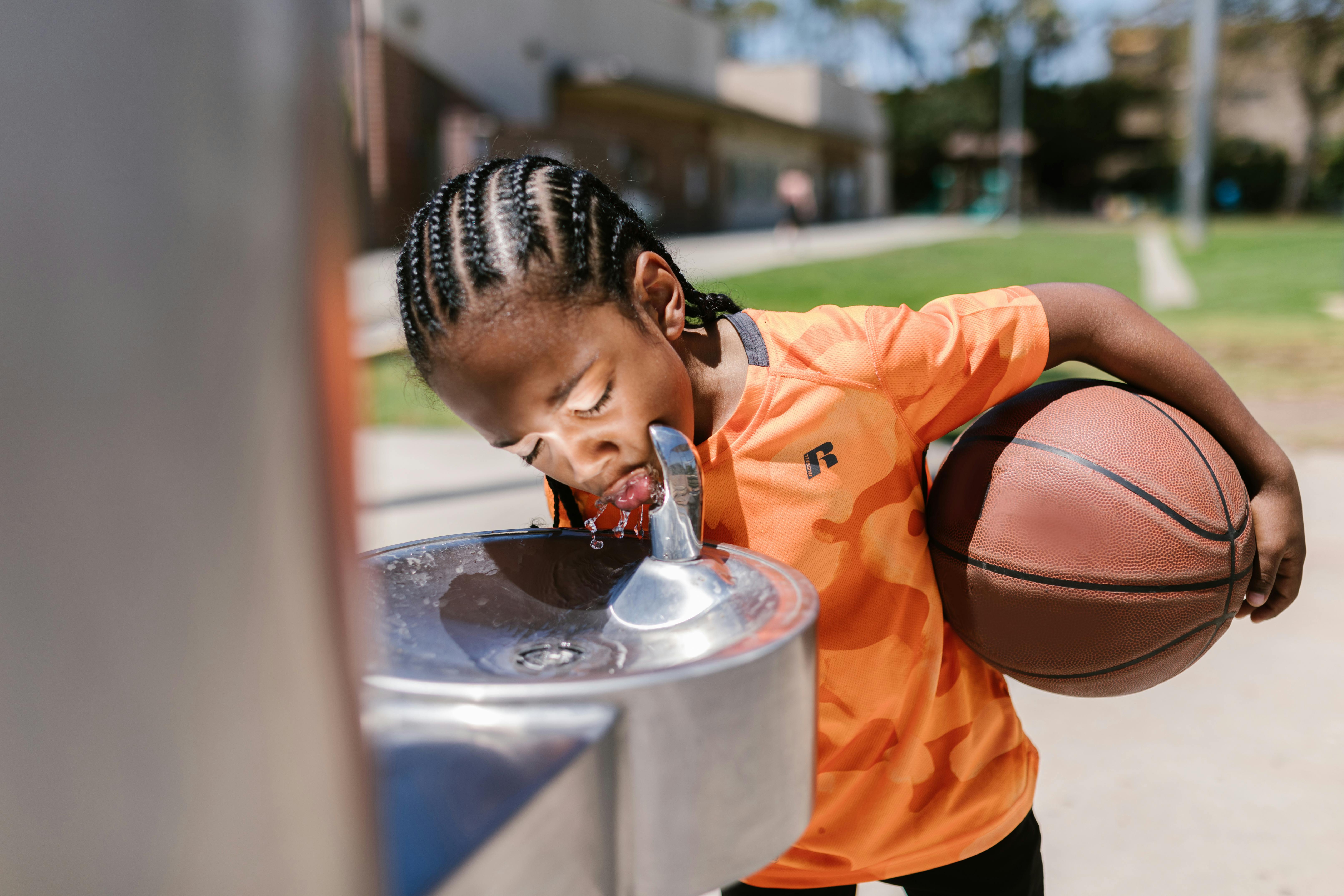 boy holding a ball while drinking from water fountain