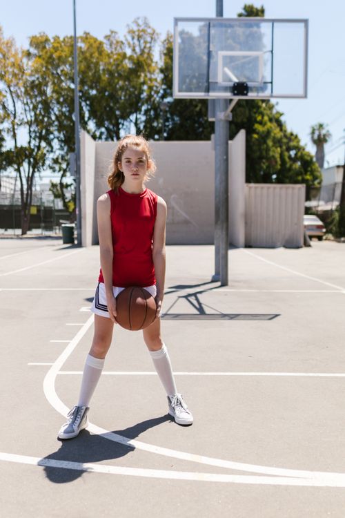 Free A teenage girl in athletic wear holds a basketball on an outdoor court under a sunny sky. Stock Photo
