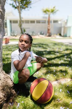 A young boy with braided hair sits by a tree with a basketball in a sunny park.
