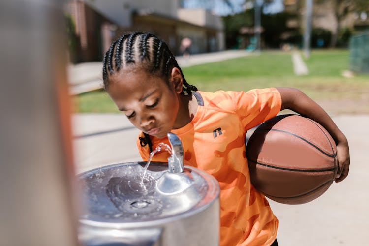 Boy Holding A Ball Drinking From A Water Fountain