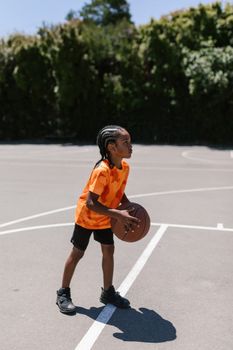A young boy in an orange shirt playing basketball on an outdoor court on a sunny day.