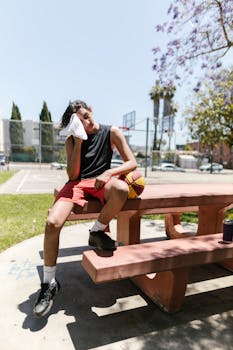 Teenager sitting on a park bench with a basketball, wiping sweat. Outdoor sports scene.