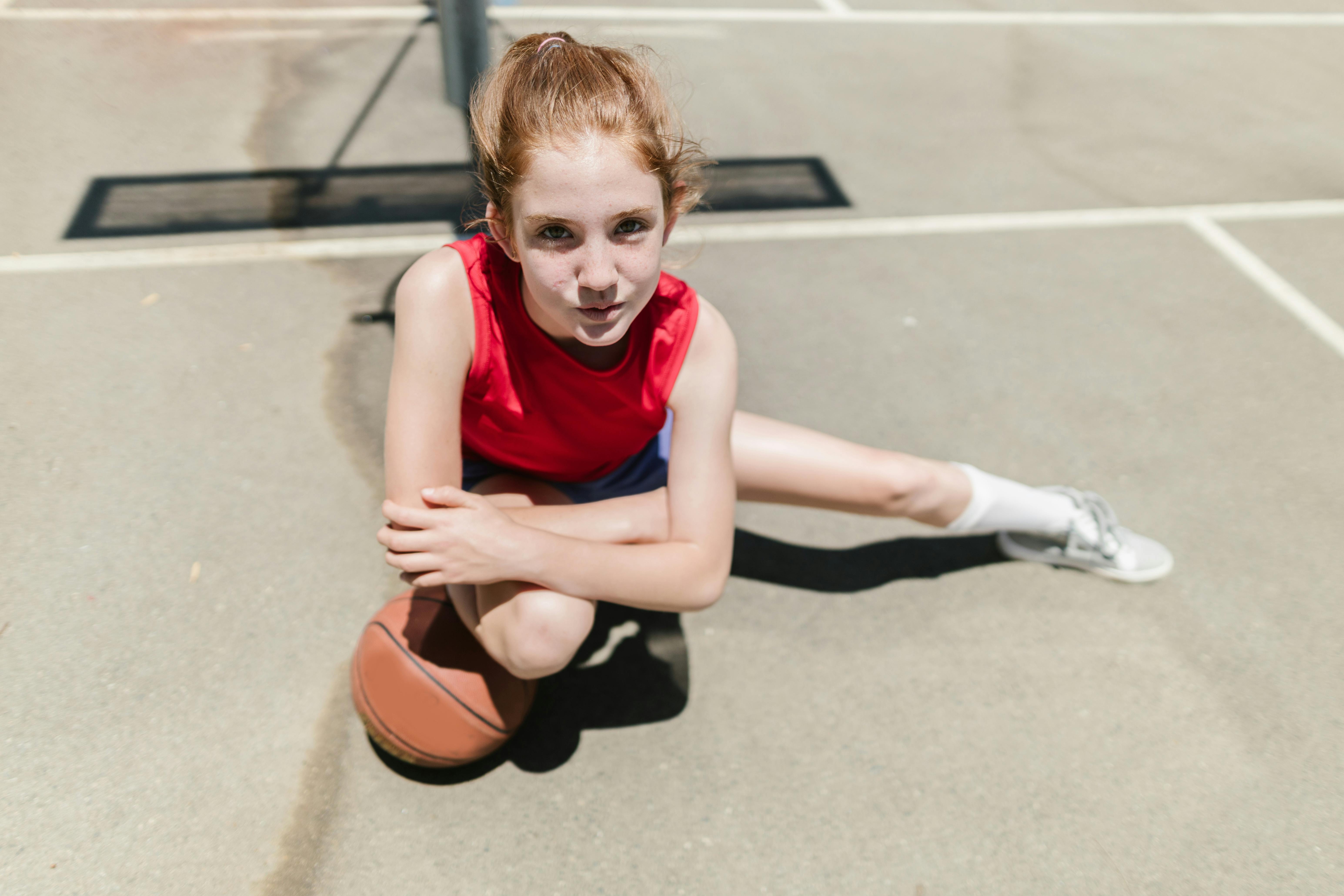 NBA photo featuring A young girl in a red tank top crouches with a basketball on an outdoor court.