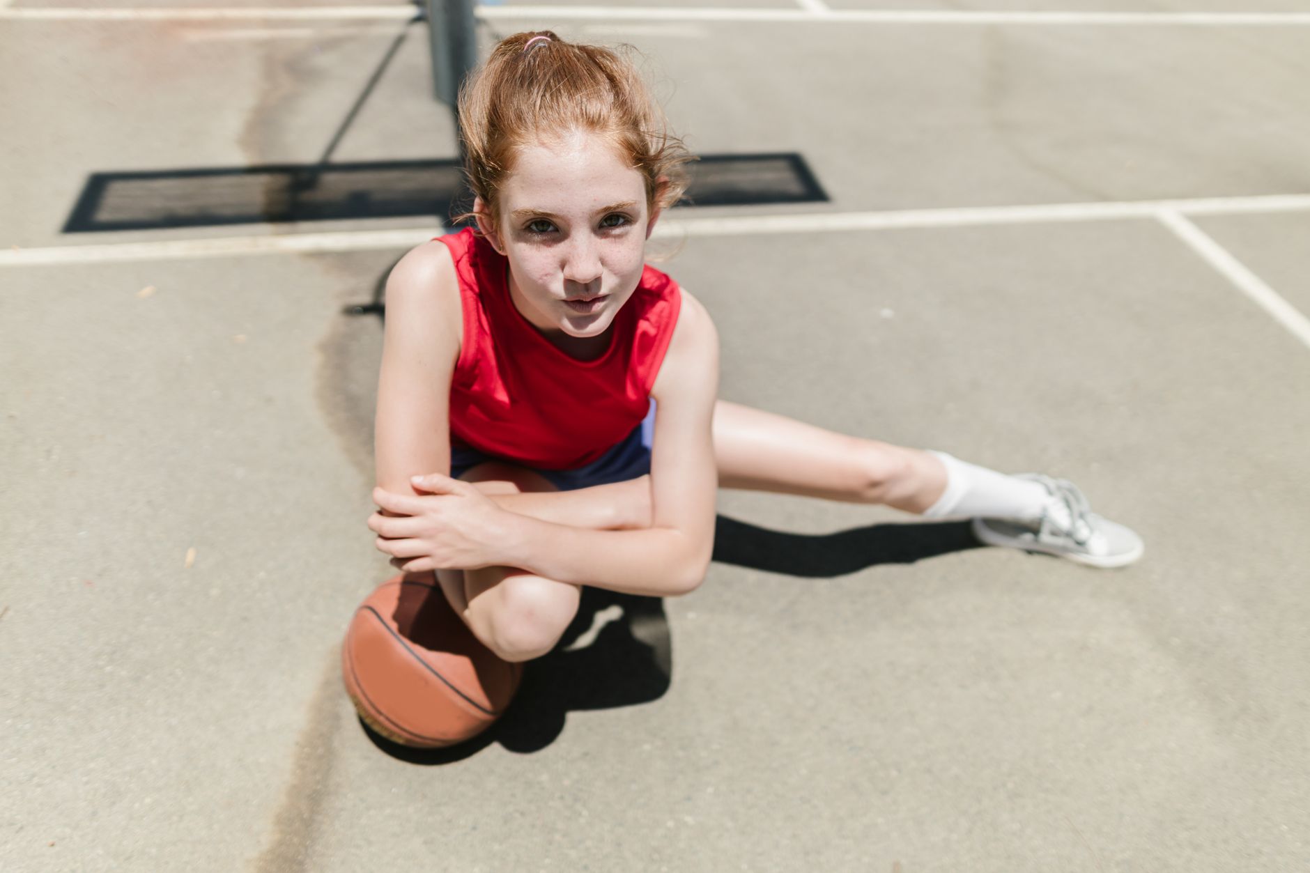 A young girl in a red tank top crouches with a basketball on an outdoor court.