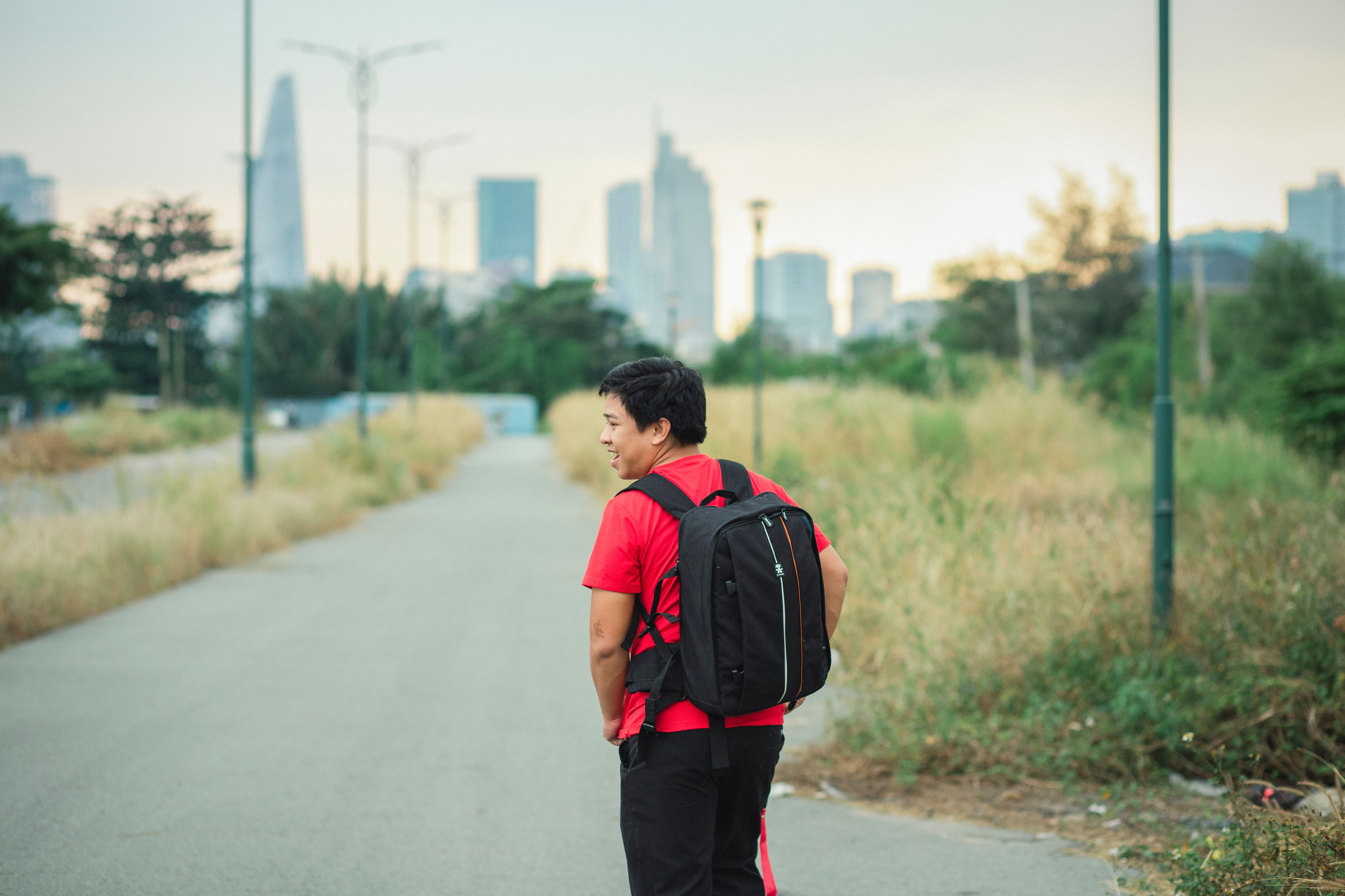 A man wearing a red shirt and backpack strolling on a path with city skyline background.
