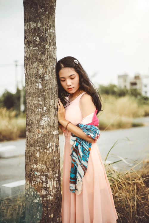 Free Elegant woman standing by a tree in a pastel dress, gazing down. Perfect for fashion and lifestyle themes. Stock Photo