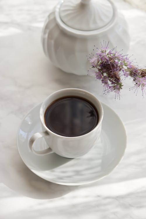 Free White ceramic cup of coffee on saucer placed near white ceramic sugar bowl and fresh flower on light background Stock Photo