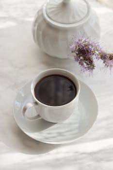White ceramic cup of coffee on saucer placed near white ceramic sugar bowl and fresh flower on light background