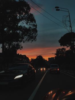 Cars drive on a silhouette-lined urban road under a vibrant sunset sky.