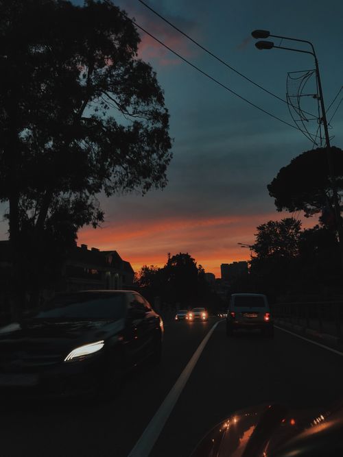 Free Cars drive on a silhouette-lined urban road under a vibrant sunset sky. Stock Photo