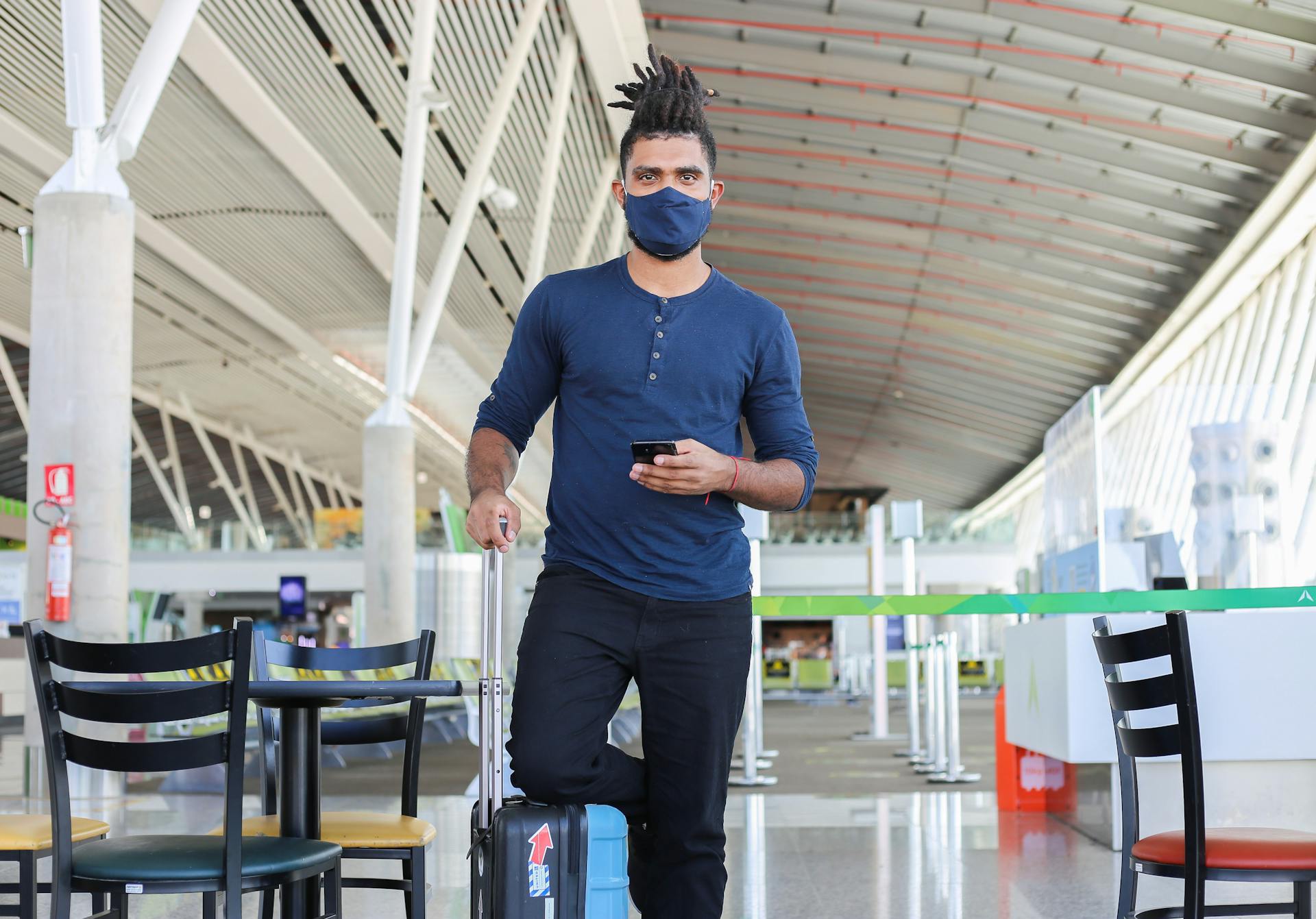 Young man wearing a face mask, holding a smartphone, and pulling luggage in a modern airport terminal.