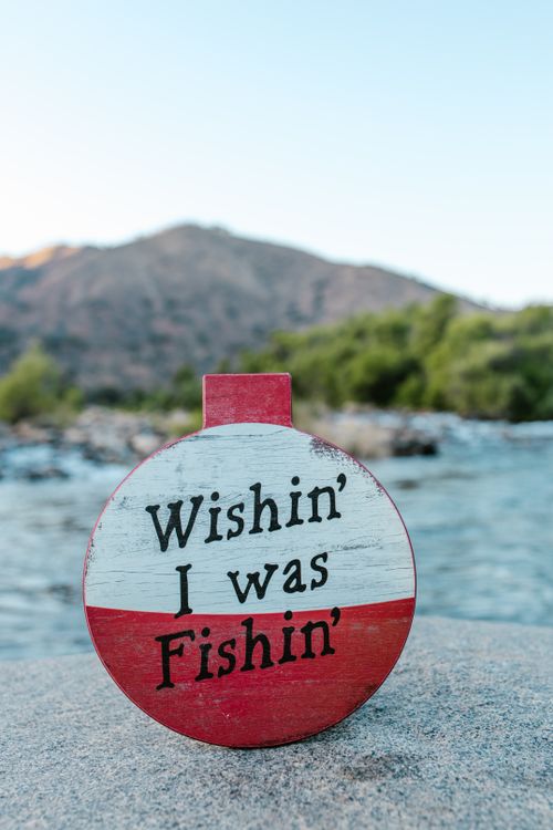 Free A fishing-themed bobber with 'Wishin' I was Fishin'' text by a scenic riverside. Stock Photo