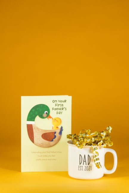 Father's Day gift featuring a mug with golden ribbons and a greeting card on a yellow background.