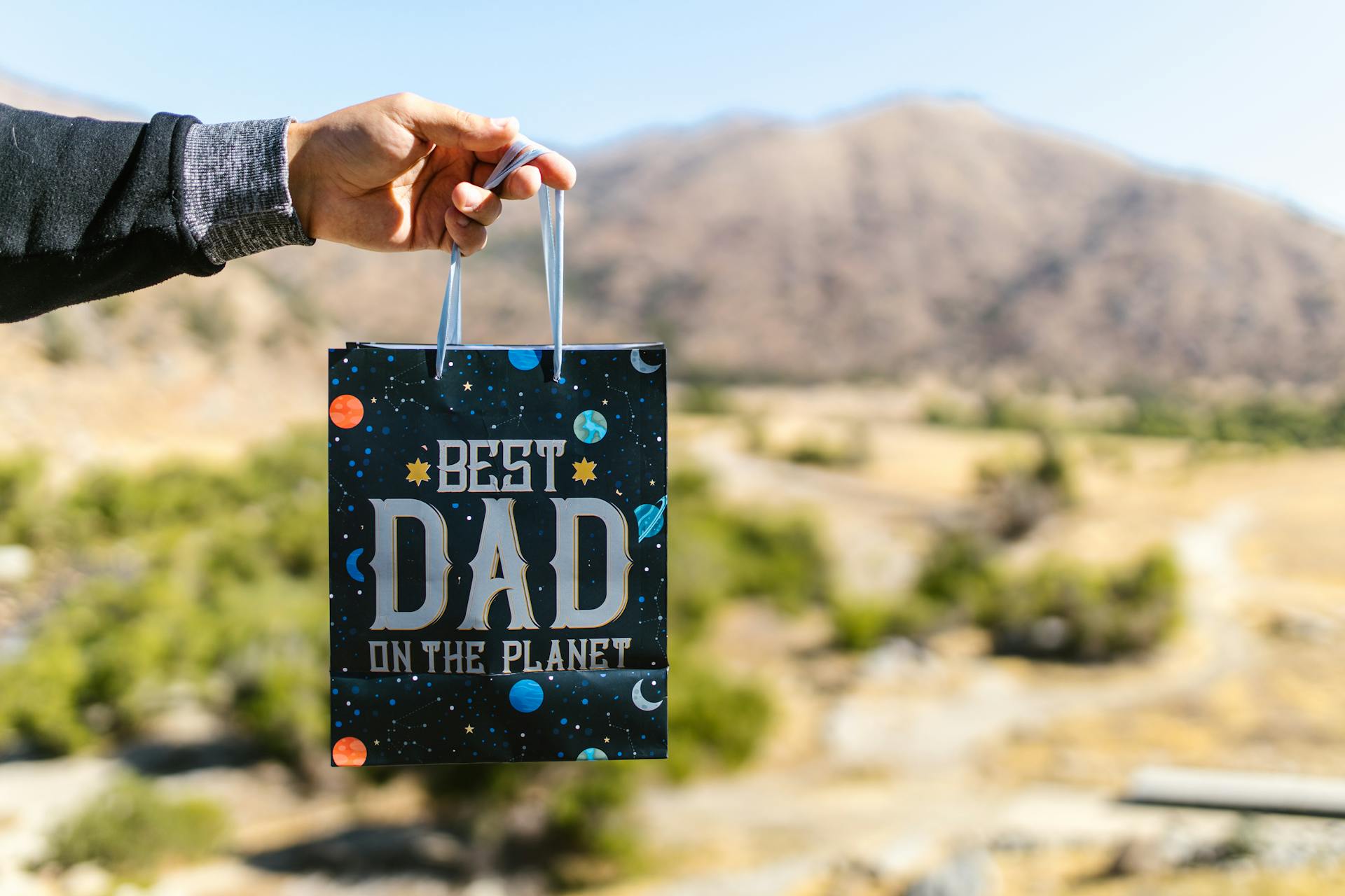 A hand holds a 'Best Dad on the Planet' gift bag outdoors, perfect for Father's Day.