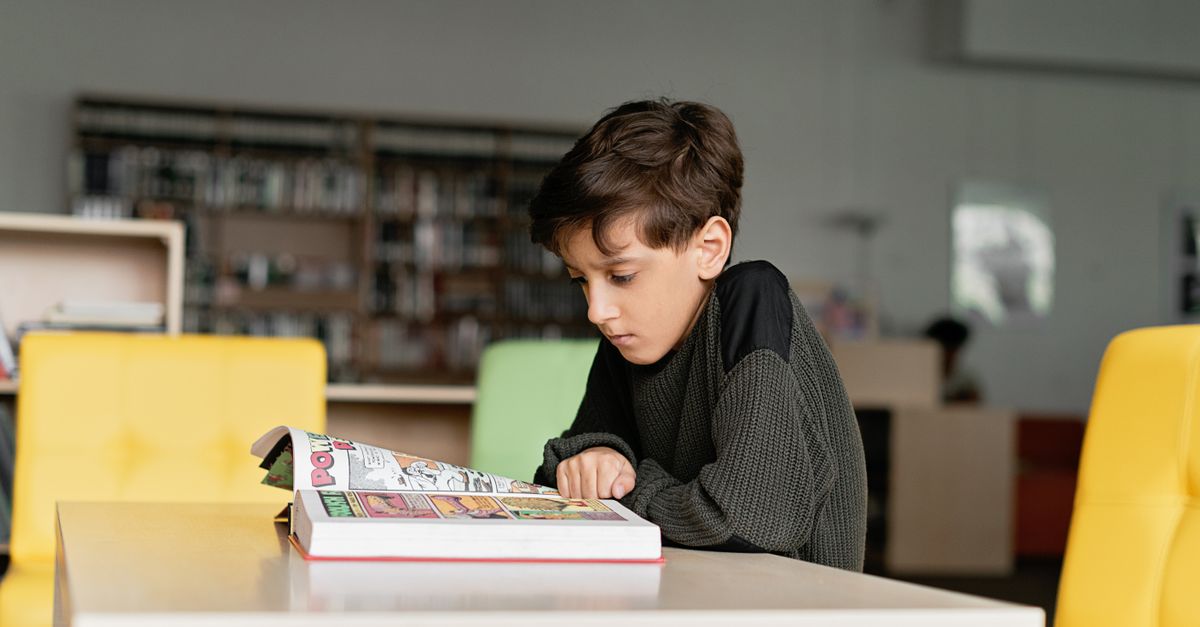 A boy in a knitted sweater reads a book attentively in a library.